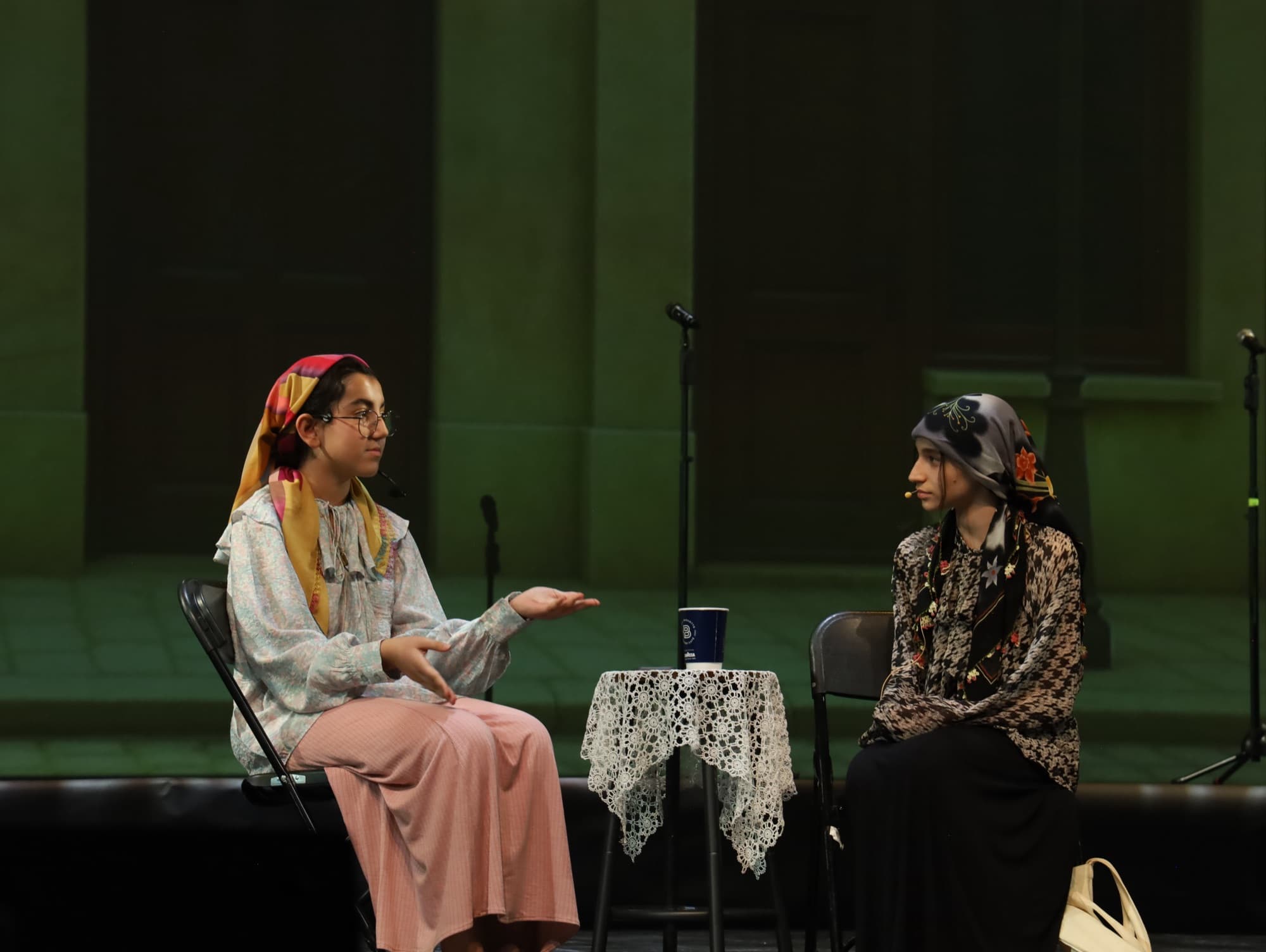 Two young performers in headscarves act a scene at a lace-covered table