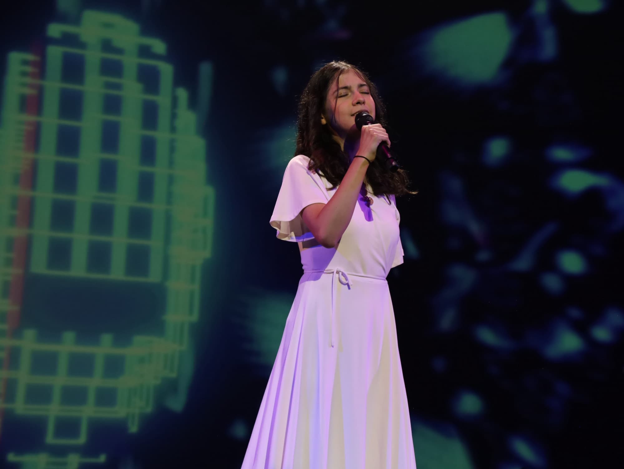 A young singer in a white dress performs under stage lights